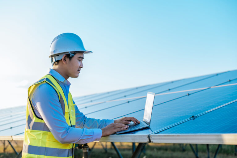 Young Asian Inspector Engineer man use laptop computer working at solar farm, Technician, supervisor male in white helmet Checking operation of sun and photovoltaic solar panel in station, copy space