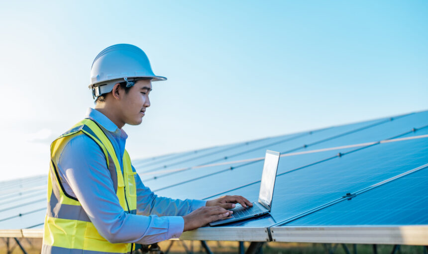 Young Asian Inspector Engineer man use laptop computer working at solar farm, Technician, supervisor male in white helmet Checking operation of sun and photovoltaic solar panel in station, copy space
